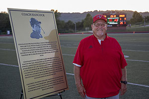 Man in red shirt and ballcap in front of sign.
